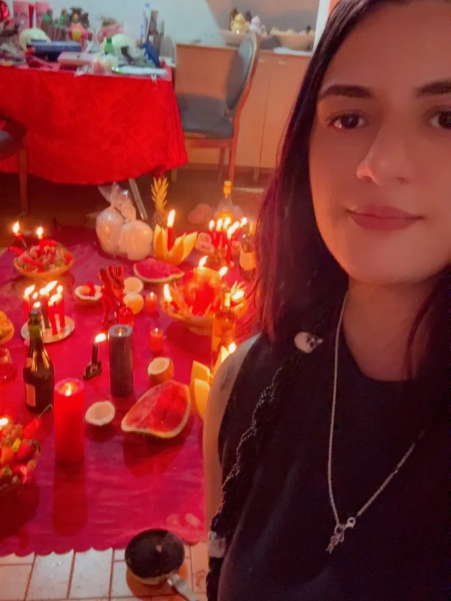 Mentora Kamily ao lado de um altar com velas vermelhas, frutas e oferendas sobre pano vermelho